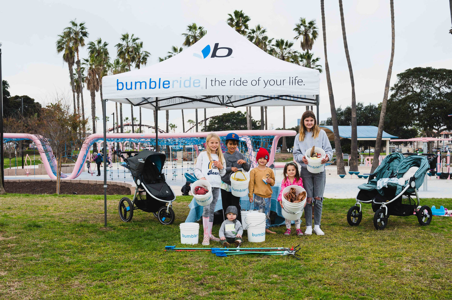 Picture of Bumbleride Park Cleanup Kids holding buckets angled to show their haul. Standing in front of bumbleride pop up tent on grass with park in background.