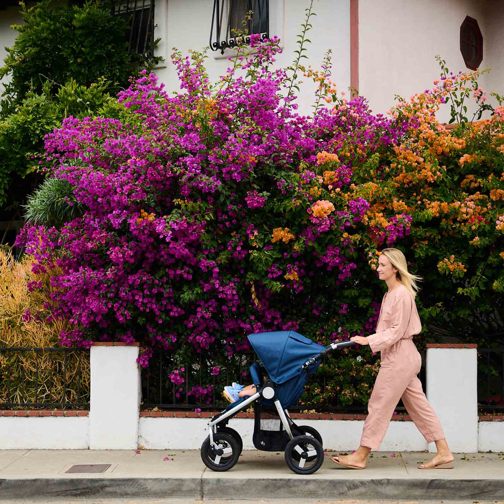 
                  
                    Mother pushing Bumbleride Indie Twin double stroller in Maritime in front of bright colored flowers on sidewalk. Global
                  
                