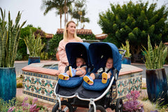 Picture of mother sitting at tiled fountain with Bumbleride Indie Twin stroller with twins with surrounding potted snake plants and palm trees in background. Global