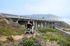 Picture of mother pushing Bumbleride Era reversible stroller on dirt trail along coast with ocean and train tracks in background. New Collection 2022 - Global