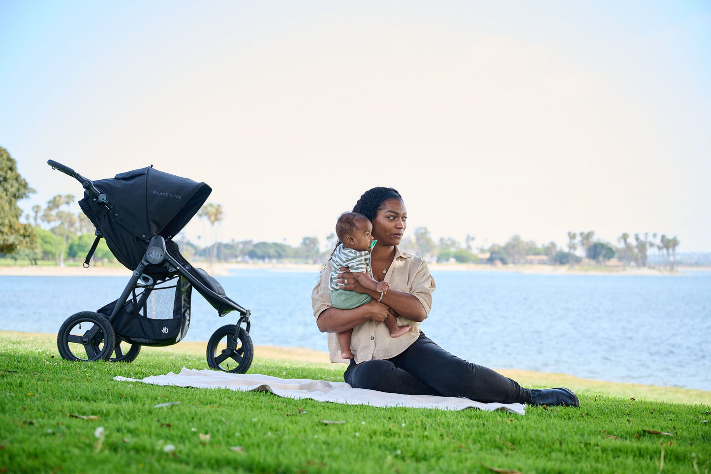 Mother holding child to chest sitting on white blanket on grass in front of the bay with Bumbleride Indie in Black parked next to them- New Collection 2022 - Global