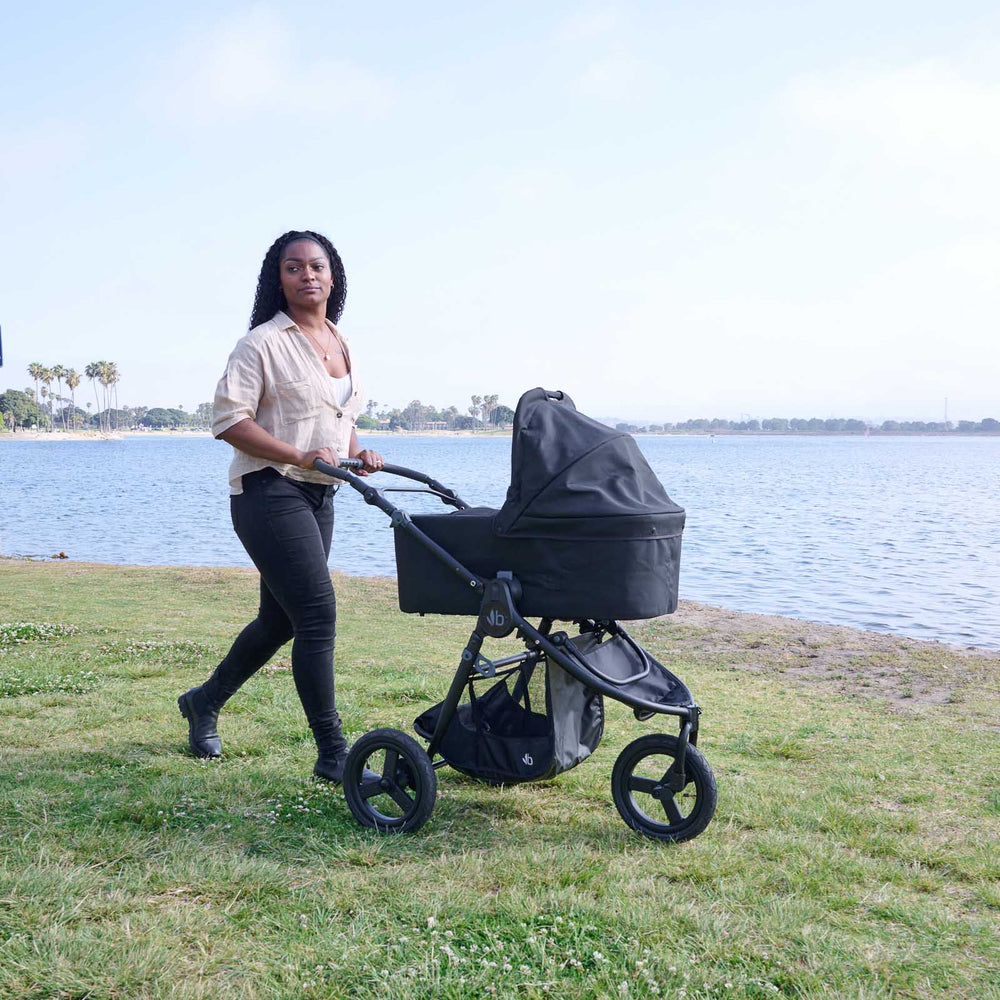 
                  
                    Bumbleride Single Bassinet in Black attached on Bumbleride Indie All Terrain Stroller in Black on grass near water with mother pushing from behind stroller. Global
                  
                