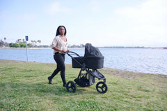 Bumbleride Single Bassinet in Black attached on Bumbleride Indie All Terrain Stroller in Black on grass near water with mother pushing from behind stroller. Global