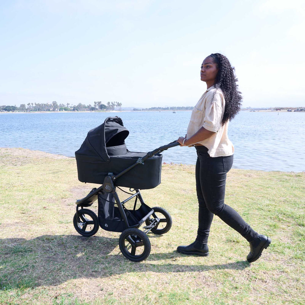 
                  
                    Mother looking into the distance pushing a Bumbleride Indie All Terrain Stroller with Bassinet in Black attached on grass with bay in background. Global
                  
                