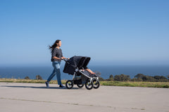 Picture of mother pushing Bumbleride Indie Twin double stroller in dusk on concrete in front of ocean and trees in background. Global