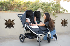 Picture of mother kneeling next to Bumbleride Indie Twin double stroller in Dusk with two children sitting in stroller. Global