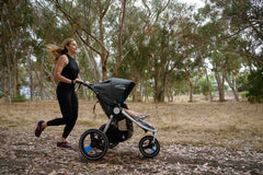 Picture of mother running a dirt trail  with toddler in an IRONMAN stroller by Bumbleride with trees in background