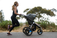Mother running with IRONMAN jogging stroller by Bumbleride with trees in background, profile view.