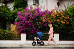 Mother pushing Bumbleride Indie Twin double stroller in Maritime in front of bright colored flowers on sidewalk. Global